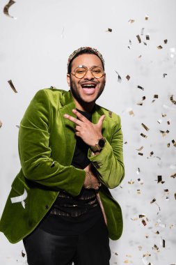 positive indian hip hop performer in green velvet blazer and crown near falling confetti on grey