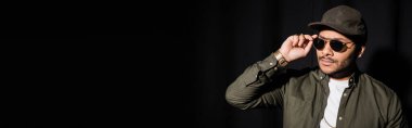 eastern hip hop performer in cap adjusting sunglasses on black, banner