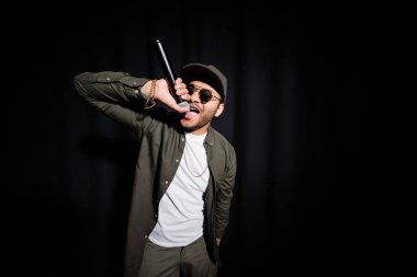 middle east hip hop performer in sunglasses and cap singing in microphone on black