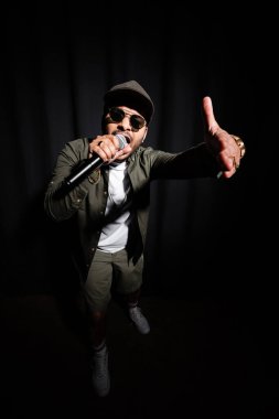 emotional middle east hip hop performer in sunglasses and cap singing in microphone and gesturing on black clipart