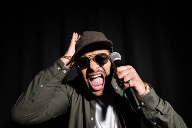emotional middle east hip hop performer in sunglasses and cap screaming while holding microphone isolated on black