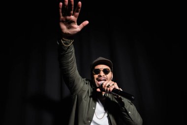 emotional middle east hip hop performer with raised hand singing in microphone on black