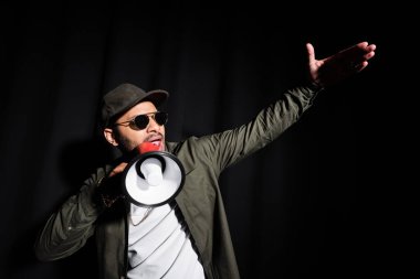 emotional middle east hip hop performer in sunglasses and cap talking in loudspeaker on black clipart