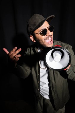 Duygusal Ortadoğu hip hop sanatçısı güneş gözlüklü ve şapkalı siyahta hoparlörden bağırıyor.
