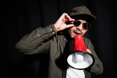 Duygusal Ortadoğu hip hop sanatçısı güneş gözlüğünü ayarlıyor ve siyahta hoparlörle konuşuyor.