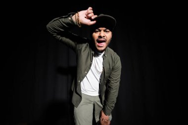 emotional and stylish indian hip hop singer adjusting cap while singing on black  clipart