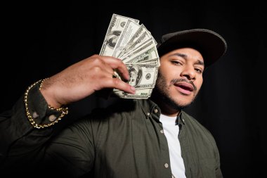 bearded indian hip hop singer in cap holding dollar banknotes isolated on black 