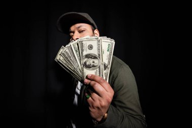 bearded indian hip hop performer in cap covering face with dollar banknotes on black 