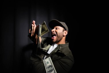 amazed indian hip hop performer in cap throwing dollar banknotes on black 