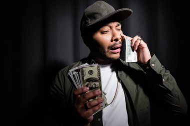 sad indian hip hop performer in cap crying and wiping tear with dollar banknote on black 