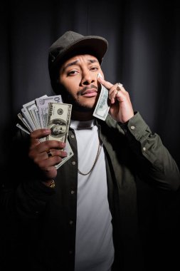 sad indian hip hop performer in cap wiping tear with dollar banknote on black 
