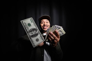 rich indian hip hop performer in cap throwing dollar banknotes and looking at camera on black 
