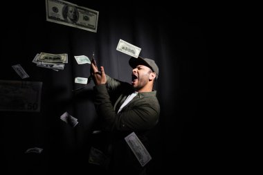 excited eastern hip hop performer in cap throwing dollar banknotes on black 