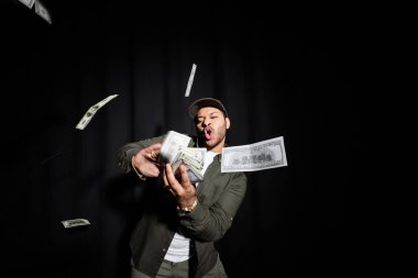 wealthy indian hip hop performer in cap throwing blurred dollar banknotes on black 