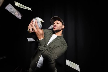 rich indian hip hop performer in cap throwing blurred dollar banknotes on black 