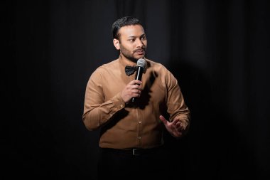 eastern comedian in shirt and bow tie holding microphone during monologue on black  clipart