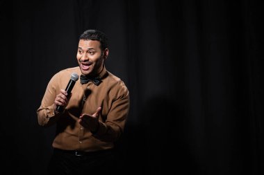 positive indian comedian in shirt and bow tie holding microphone during monologue on black 