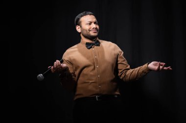 skeptical indian comedian in shirt and bow tie gesturing while holding microphone during monologue on black  clipart