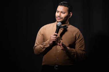 skeptical indian comedian in shirt and bow tie holding microphone during monologue on black  clipart