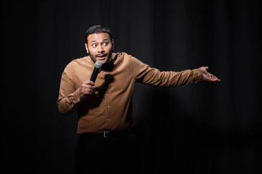 puzzled indian comedian in shirt and bow tie holding microphone and gesturing during monologue on black  clipart