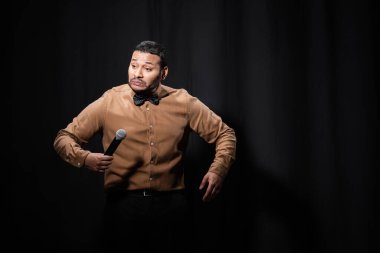 indian stand up comedian grimacing while telling joke into microphone on dark stage on black 
