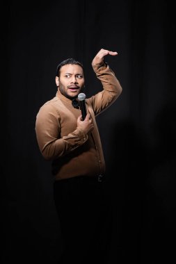 indian stand up comedian gesturing while telling joke into microphone on dark stage on black  clipart