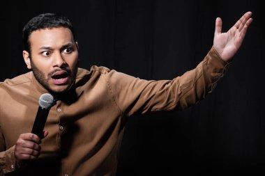 shocked indian stand up comedian telling joke into microphone on dark stage isolated on black 