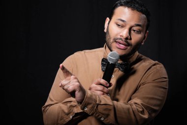 indian stand up comedian pointing with finger while telling jokes into microphone isolated on black 