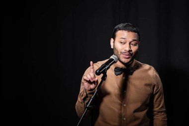 indian stand up comedian pointing with finger while telling jokes into microphone on stand on black background clipart