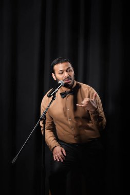 Hintli komedyen stand-up komedisi yapıyor ve mikrofonda siyah üzerine şakalar yapıyor. 