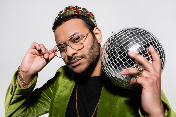 eastern hip hop performer in green velvet blazer and crown holding disco ball while adjusting eyeglasses isolated on grey