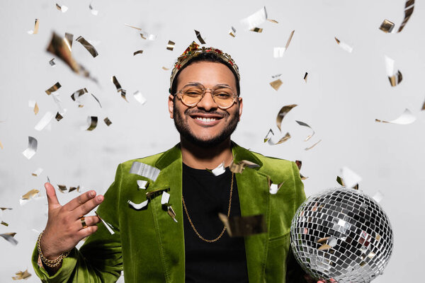 happy indian hip hop performer in green velvet blazer and crown holding disco ball near falling confetti on grey