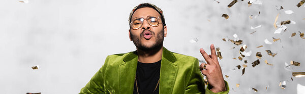 indian hip hop performer in crown showing peace near falling confetti on grey, banner