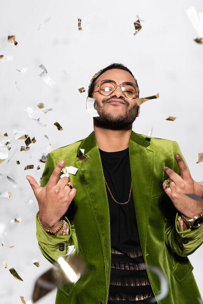 confident indian hip hop performer in green velvet blazer and crown near falling confetti on grey