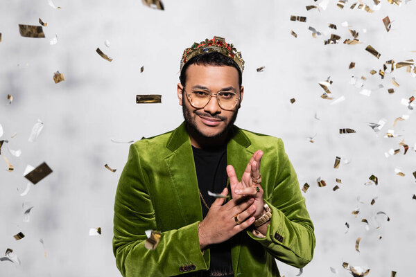 pleased indian hip hop performer in green velvet blazer and crown near falling confetti on grey