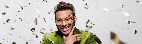 cheerful indian hip hop performer in green velvet blazer and crown near falling confetti on grey, banner