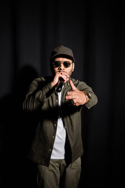 eastern hip hop performer in sunglasses singing in microphone and pointing at camera on black
