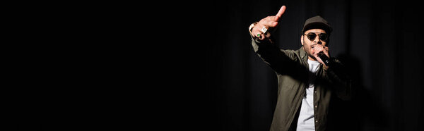 indian hip hop performer in sunglasses singing in microphone on black, banner