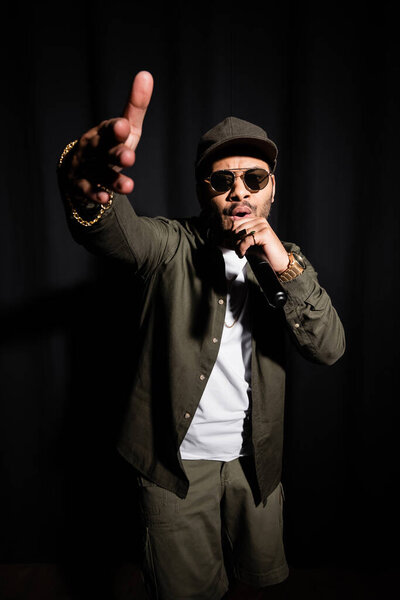 eastern hip hop performer in sunglasses gesturing while singing in microphone on black