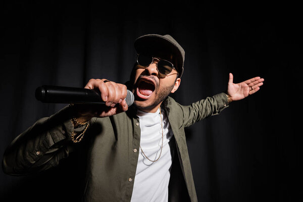 emotional eastern hip hop performer in sunglasses singing in microphone on black