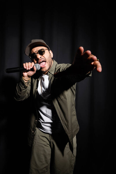 eastern hip hop performer in sunglasses with open mouth singing in microphone on black