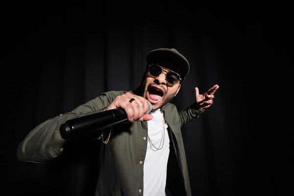 indian hip hop performer in sunglasses and cap singing in microphone on black