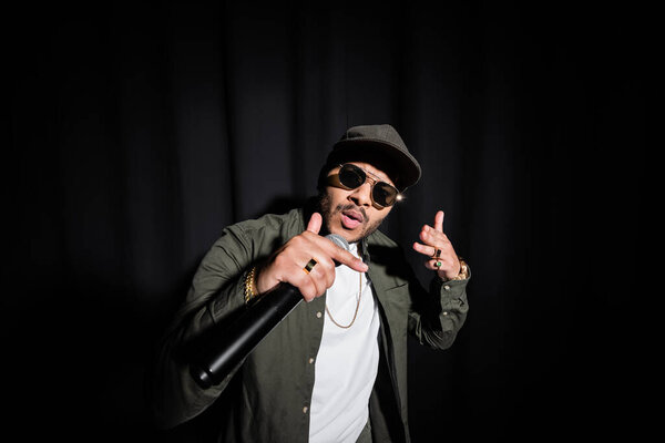 indian hip hop singer in sunglasses and cap singing in microphone on black