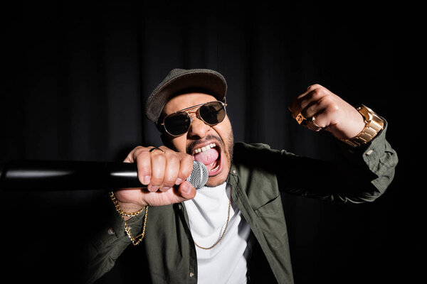indian hip hop performer in sunglasses and cap singing loud in microphone on black