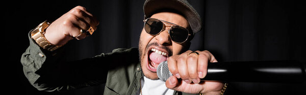 indian hip hop performer in sunglasses and cap singing loud in microphone on black, banner
