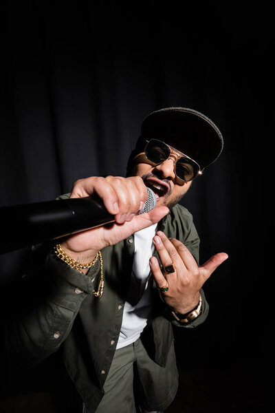 indian hip hop performer in sunglasses singing in microphone on black