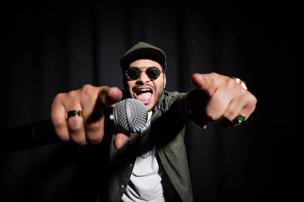 excited middle east hip hop performer in sunglasses and cap singing in microphone and gesturing on black