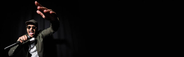 emotional indian hip hop performer with raised hand singing in microphone on black, banner