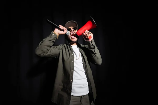 emotional indian hip hop singer in sunglasses and cap screaming in microphone and loudspeaker on black