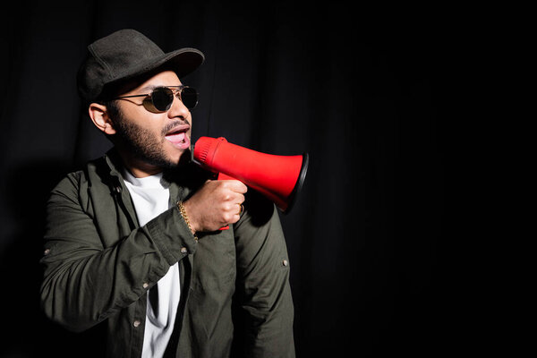 emotional indian hip hop performer in sunglasses and cap singing and holding loudspeaker on black